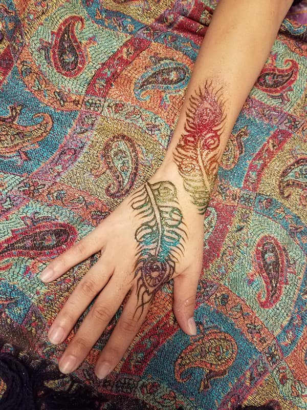 Two glitter henna peacock feathers in rainbow colours across a hand and forearm